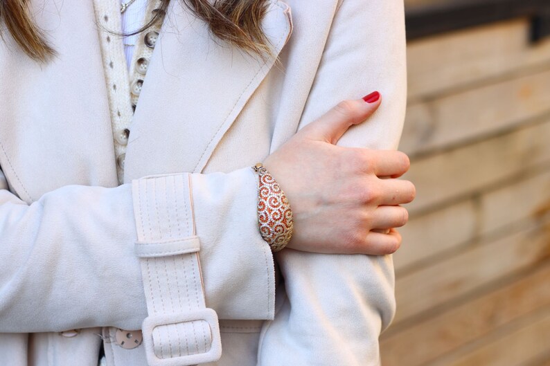 K&ouml;nnte beinhalten: Nahaufnahme eines Frauenhandgelenks, das ein gold- und orangefarbenes Armband mit einem Spiralmuster tr&auml;gt. Das Armband ist aus Metall und hat ein gro&szlig;es, ovales Design. Die Frau tr&auml;gt einen wei&szlig;en Mantel und hat roten Nagellack.