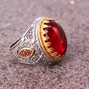 May include: A silver ring with a large red gemstone set in a gold bezel. The ring is decorated with intricate silver and gold designs and small red gemstones.