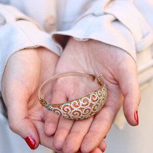 K&ouml;nnte beinhalten: Ein goldenes Armreif-Armband mit einem Spiralmuster und orangefarbenen und wei&szlig;en Steinen.