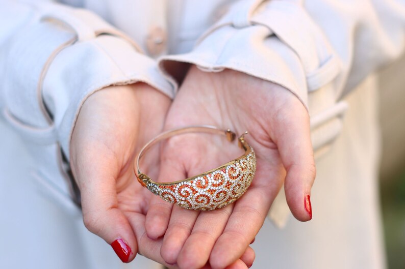 K&ouml;nnte beinhalten: Ein goldenes Armreif-Armband mit einem Spiralmuster und orangefarbenen und wei&szlig;en Steinen.