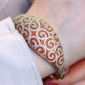 Könnte beinhalten: Ein goldenes Armreif-Armband mit einem Wirbeldesign, besetzt mit orangefarbenen und weißen Steinen.