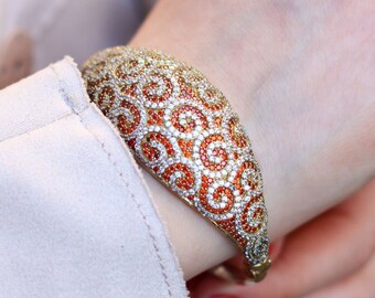 May include: A gold bangle bracelet with a swirl design set with orange and white stones.