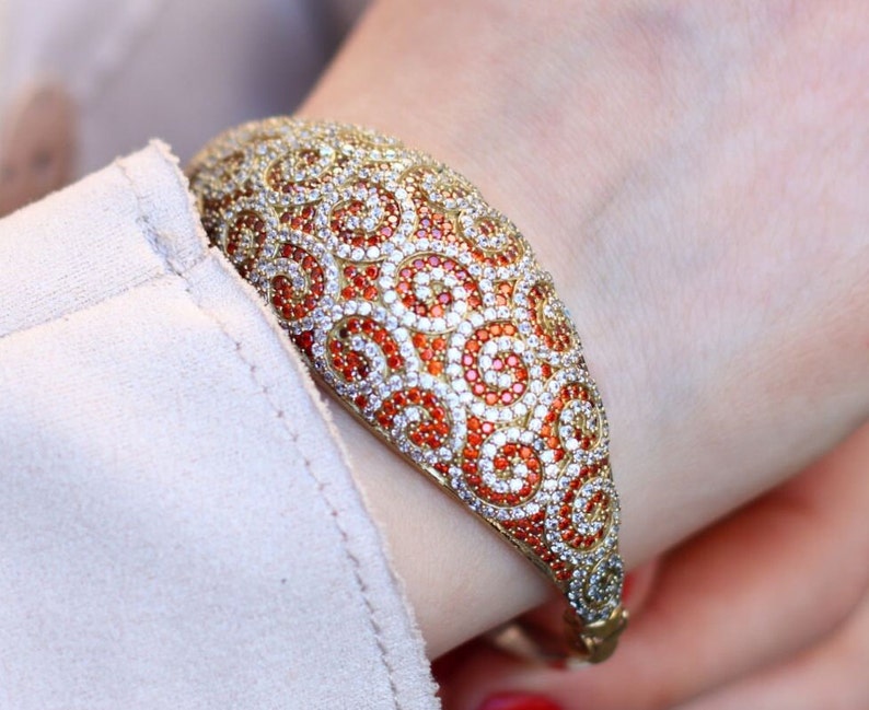 K&ouml;nnte beinhalten: Ein goldenes Armreif-Armband mit einem Wirbeldesign, besetzt mit orangefarbenen und wei&szlig;en Steinen.