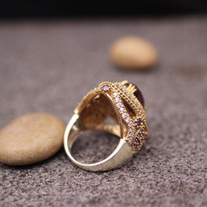 Puede incluir: Un anillo de oro con una gran piedra preciosa p&uacute;rpura rodeada de piedras m&aacute;s peque&ntilde;as transparentes y p&uacute;rpuras. El anillo est&aacute; colocado sobre una superficie marr&oacute;n texturizada.