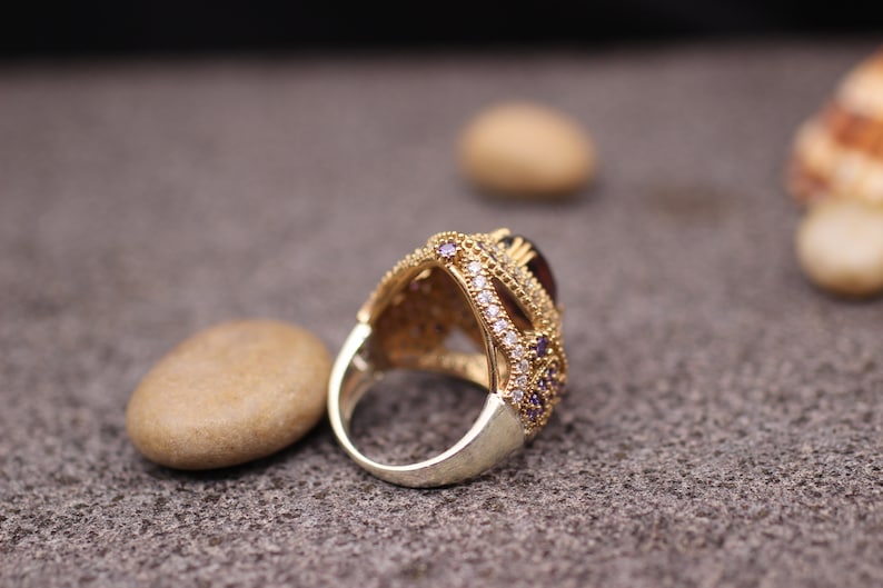 Puede incluir: Un anillo de oro con una gran piedra preciosa p&uacute;rpura rodeada de piedras m&aacute;s peque&ntilde;as transparentes y p&uacute;rpuras. El anillo est&aacute; colocado sobre una superficie marr&oacute;n texturizada.