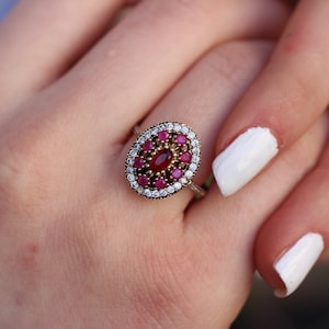 May include: A silver ring featuring an oval red gemstone surrounded by smaller red gemstones and clear cubic zirconia stones.