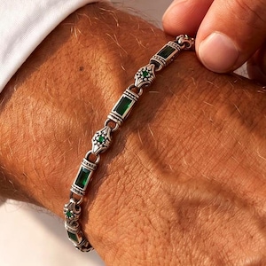 Può includere: Un bracciale a maglie color argento caratterizzato dall'alternanza di pietre verdi rettangolari e piccoli segmenti metallici decorati con pietre verdi rotonde incastonate. Il design presenta un motivo ripetitivo di queste maglie.