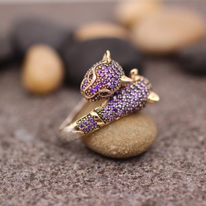K&ouml;nnte beinhalten: Ein gold-silberner Ring mit zwei Pantherk&ouml;pfen, besetzt mit lila Edelsteinen. Der Ring liegt auf einem kleinen Stein, mit einem verschwommenen Hintergrund aus anderen Steinen. Ein Schmuckst&uuml;ck.