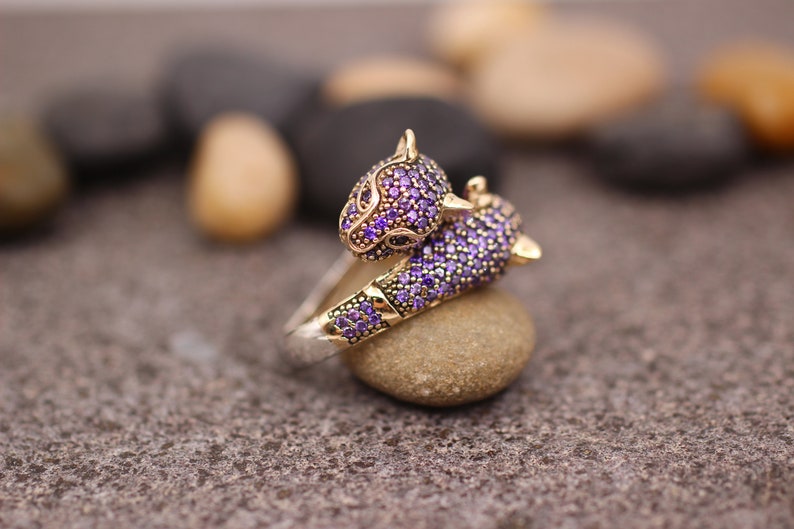 K&ouml;nnte beinhalten: Ein gold-silberner Ring mit zwei Pantherk&ouml;pfen, besetzt mit lila Edelsteinen. Der Ring liegt auf einem kleinen Stein, mit einem verschwommenen Hintergrund aus anderen Steinen. Ein Schmuckst&uuml;ck.