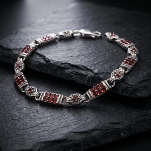 Könnte beinhalten: Ein silbernes Kettenarmband mit abwechselnden rechteckigen Gliedern, die mit kleinen quadratischen roten Steinen besetzt sind, und verzierten runden Gliedern, die einen einzelnen runden roten Stein enthalten. Das Metall hat ein dunkles Finish.