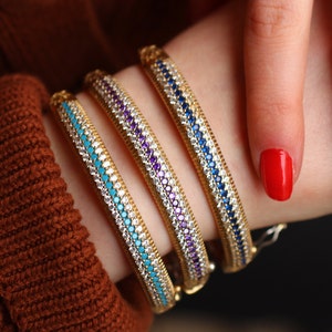 May include: Three gold bangle bracelets with a row of turquoise, purple, and blue gemstones.