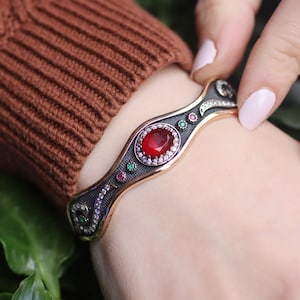 May include: Ornate silver and gold bracelet with a central oval red gemstone, surrounded by small white, green, and pink stones. The bracelet has a detailed, patterned design and is shown on a wrist.
