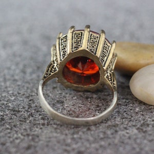 Peut inclure: Une bague en or avec un motif orn&eacute; et une grosse pierre pr&eacute;cieuse rouge. La bague est pos&eacute;e sur une surface grise textur&eacute;e.
