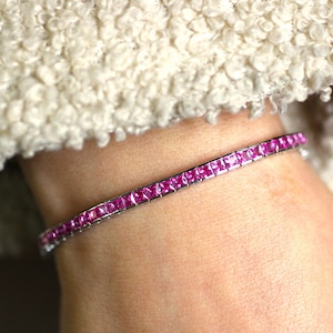 Può includere: Un bracciale d'argento con una fila di pietre preziose rosa vivo e quadrate. Il bracciale è indossato su un polso, con un tessuto sfocato color crema sullo sfondo. Le pietre sono incastonate vicine, creando una linea continua di colore.