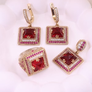 Puede incluir: Un conjunto de joyas doradas con detalles de piedras preciosas rojas. El conjunto incluye un anillo, un par de pendientes y un colgante. Las joyas están decoradas con pequeñas piedras transparentes.