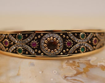 Puede incluir: Un brazalete de oro con un intrincado diseño de filigrana en blanco y negro. El brazalete está adornado con piedras preciosas verdes y rojas.