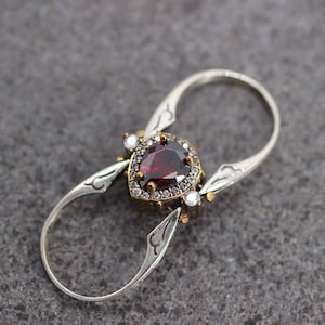 Op de afbeelding: Een zilveren ring met een rode edelsteen omringd door kleinere witte edelstenen. De ring heeft een uniek dubbelband ontwerp.