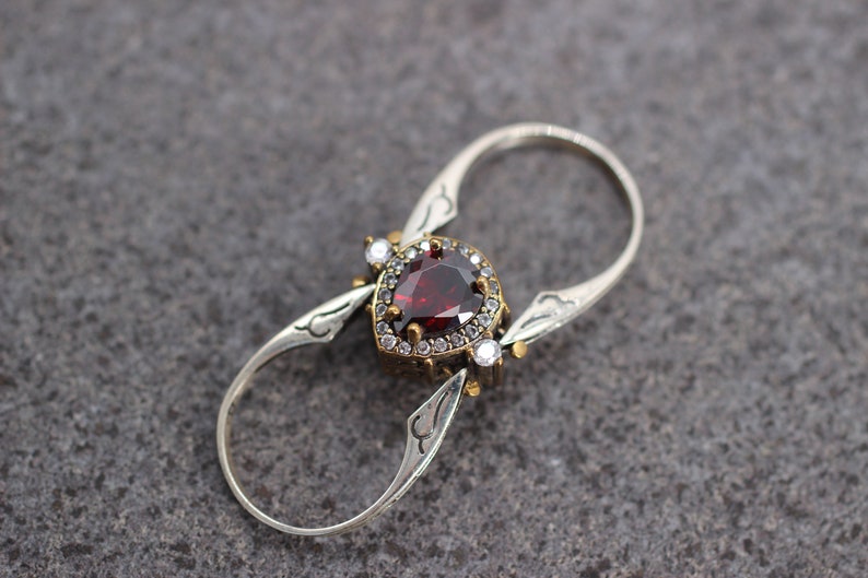 Op de afbeelding: Een zilveren ring met een rode edelsteen omringd door kleinere witte edelstenen. De ring heeft een uniek dubbelband ontwerp.