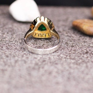 May include: A gold ring with a green gemstone set in a triangular shape. The ring is set on a gray textured surface.