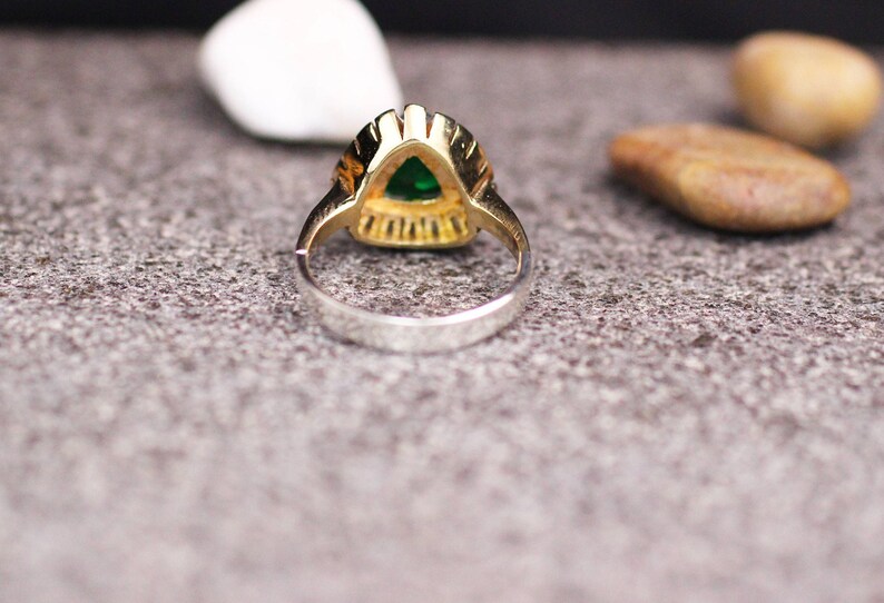 May include: A gold ring with a green gemstone set in a triangular shape. The ring is set on a gray textured surface.