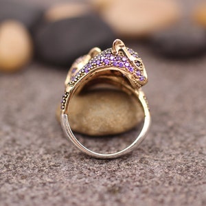 K&ouml;nnte beinhalten: Ein goldfarbener Ring mit einem Leopardenkopf-Design, verziert mit kleinen lila Edelsteinen. Der Ring hat ein silbernes Band und ist vor einem neutralen Hintergrund platziert, der die komplizierten Details des Schmucks hervorhebt.