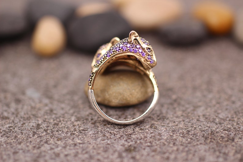 K&ouml;nnte beinhalten: Ein goldfarbener Ring mit einem Leopardenkopf-Design, verziert mit kleinen lila Edelsteinen. Der Ring hat ein silbernes Band und ist vor einem neutralen Hintergrund platziert, der die komplizierten Details des Schmucks hervorhebt.
