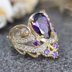 May include: A gold ring with a large pear-shaped purple gemstone surrounded by smaller purple and white gemstones.