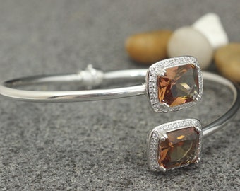 Puede incluir: Un brazalete de plata con dos engastes de piedras preciosas marrones. Cada piedra preciosa está rodeada por un halo de piedras más pequeñas y transparentes.