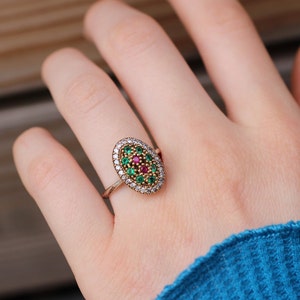 Könnte beinhalten: Ovaler Ring mit goldenem Band, besetzt mit grünen und roten Edelsteinen in der Mitte, umgeben von weißen Steinen. Der Ring wird an einer Hand mit einem blauen Ärmel getragen.
