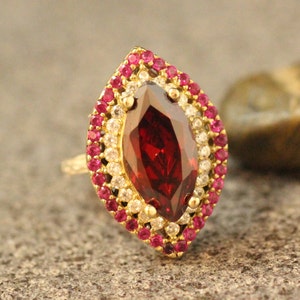 Könnte beinhalten: Ein goldener Ring mit einem großen, roten, markisenförmigen Edelstein, umgeben von einem doppelten Heiligenschein aus kleineren, klaren und roten Edelsteinen.