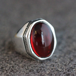 Peut inclure: Une bague en argent avec une pierre précieuse rouge sertie dans une bélière hexagonale. La bague a une bande texturée.