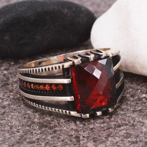 May include: A silver ring with a large, rectangular, red gemstone. The ring has three bands with small, clear stones.