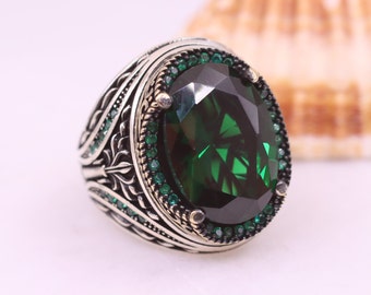 May include: A silver ring with a large, oval-shaped green gemstone surrounded by smaller green gemstones. The ring has intricate details and a silver band.