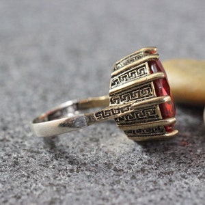 Peut inclure: Une bague en or avec une pierre pr&eacute;cieuse rouge sertie dans un motif g&eacute;om&eacute;trique. La bague est en m&eacute;tal et a une bande argent&eacute;e.