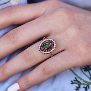 Könnte beinhalten: Ein silberner Ring mit einer ovalen Fassung, in der ein grüner Edelstein von kleineren rosa und weißen Edelsteinen umgeben ist.