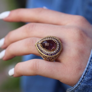 Puede incluir: Un anillo de oro con una gran piedra preciosa p&uacute;rpura en forma de pera rodeada de peque&ntilde;as piedras preciosas transparentes y p&uacute;rpuras.