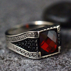 May include: A silver ring with a red gemstone and a black Greek key design.