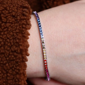Puede incluir: Una pulsera plateada con una gama de gemas cuadradas. Las gemas cambian de púrpura a rojo, creando un accesorio vibrante. La pulsera se muestra en una muñeca.