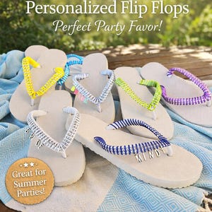 Può includere: Una collezione di infradito bianche personalizzate con cinturini colorati, tra cui giallo, blu, argento, verde e viola. Le infradito sono su un asciugamano a fantasia blu e bianco. Il testo sull'immagine recita "Personalized Flip Flops" e "Great for Summer Parties!"