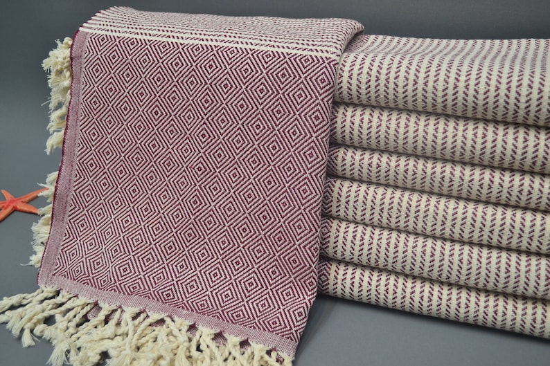 Pu&ograve; includere: Un mucchio di asciugamani turchi con un motivo a rombi di colore bordeaux e bianco. Gli asciugamani hanno frange alle estremit&agrave;.