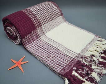 May include: A burgundy and white waffle weave Turkish towel with fringe. The towel is rolled up and has a starfish on the gray surface.