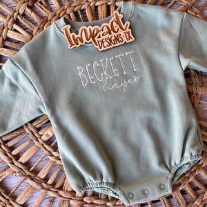 Könnte beinhalten: Ein langärmeliger Strampler in Salbeigrün mit weißem Sticktext Beckett hayes. Ein Holzschild mit der Aufschrift Impact DESIGNS TX befindet sich in der Nähe des gerippten Rundhalsausschnitts. Das Kleidungsstück hat einen Druckknopfverschluss am Saum.