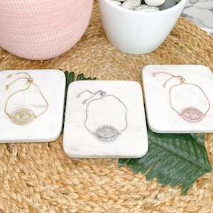Puede incluir: Tres pulseras de ojo turco ajustables sobre azulejos cuadrados blancos. Las pulseras son de oro, plata y oro rosa con detalles de cristal transparente.