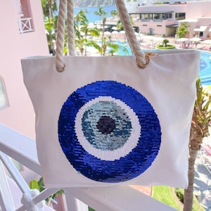 Può includere: Borsa tote in tela bianca con un grande designo di occhio turco blu fatto di paillettes. La borsa ha manici in corda.