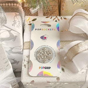 May include: A white gift box with a gold ribbon and a silver snowflake PopSocket attached to the front. The PopSocket packaging features the brand name "PopSockets" and "PopGrip".