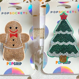 Peut inclure: Deux supports de téléphone PopSockets. L'un est un bonhomme en pain d'épices avec un nœud papillon rouge, l'autre est un arbre de Noël. Les deux sont recouverts de strass étincelants. Les PopSockets sont sur un fond blanc et irisé.