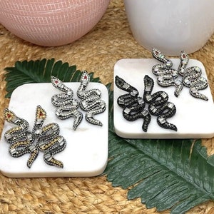 May include: Two pairs of snake-shaped earrings on white marble coasters. One pair is silver with clear rhinestones and the other is black with black rhinestones.
