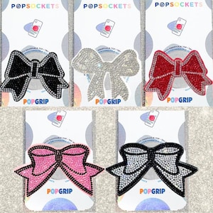 May include: A collection of PopSockets featuring bow designs. The PopSockets are available in black, silver, red, pink, and black and silver. Each bow is embellished with rhinestones and the word "POPGRIP" is printed on the bottom.