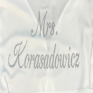 May include: A white satin robe with the text "Mrs. Korasadowicz" in silver glitter script. The robe has a V-neck design and a matching belt. The image is a close-up of the robe's upper portion.