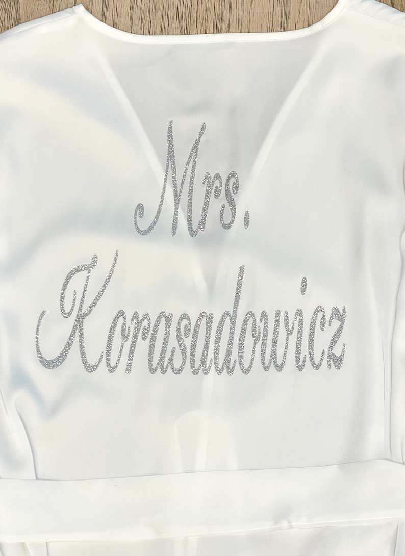 May include: A white satin robe with the text "Mrs. Korasadowicz" in silver glitter script. The robe has a V-neck design and a matching belt. The image is a close-up of the robe's upper portion.